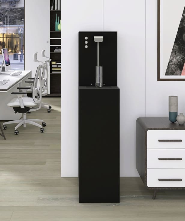 Erogatore d'acqua frizzante a colonna di colore nero, collocato in un moderno ufficio open space con arredi eleganti. L'erogatore si integra perfettamente nell'ambiente, accanto a una scrivania bianca con sedie ergonomiche e a un mobile basso con cassetti.